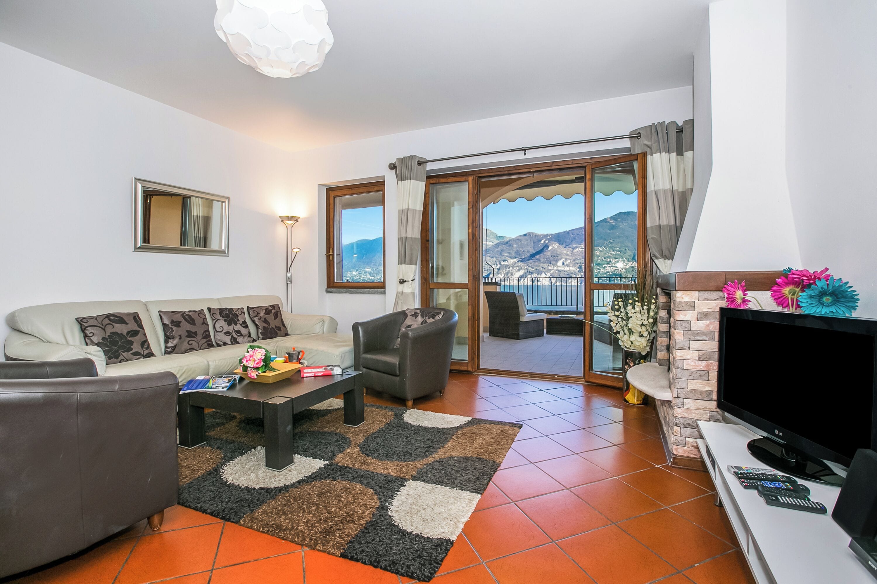 A living room with a sofa, armchair, coffee table, and TV. There is a balcony with a view of mountains.
