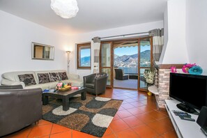 A living room with a sofa, armchair, coffee table, TV, and a view of mountains through the sliding glass door. - Bellavista Grande (Menaggio)