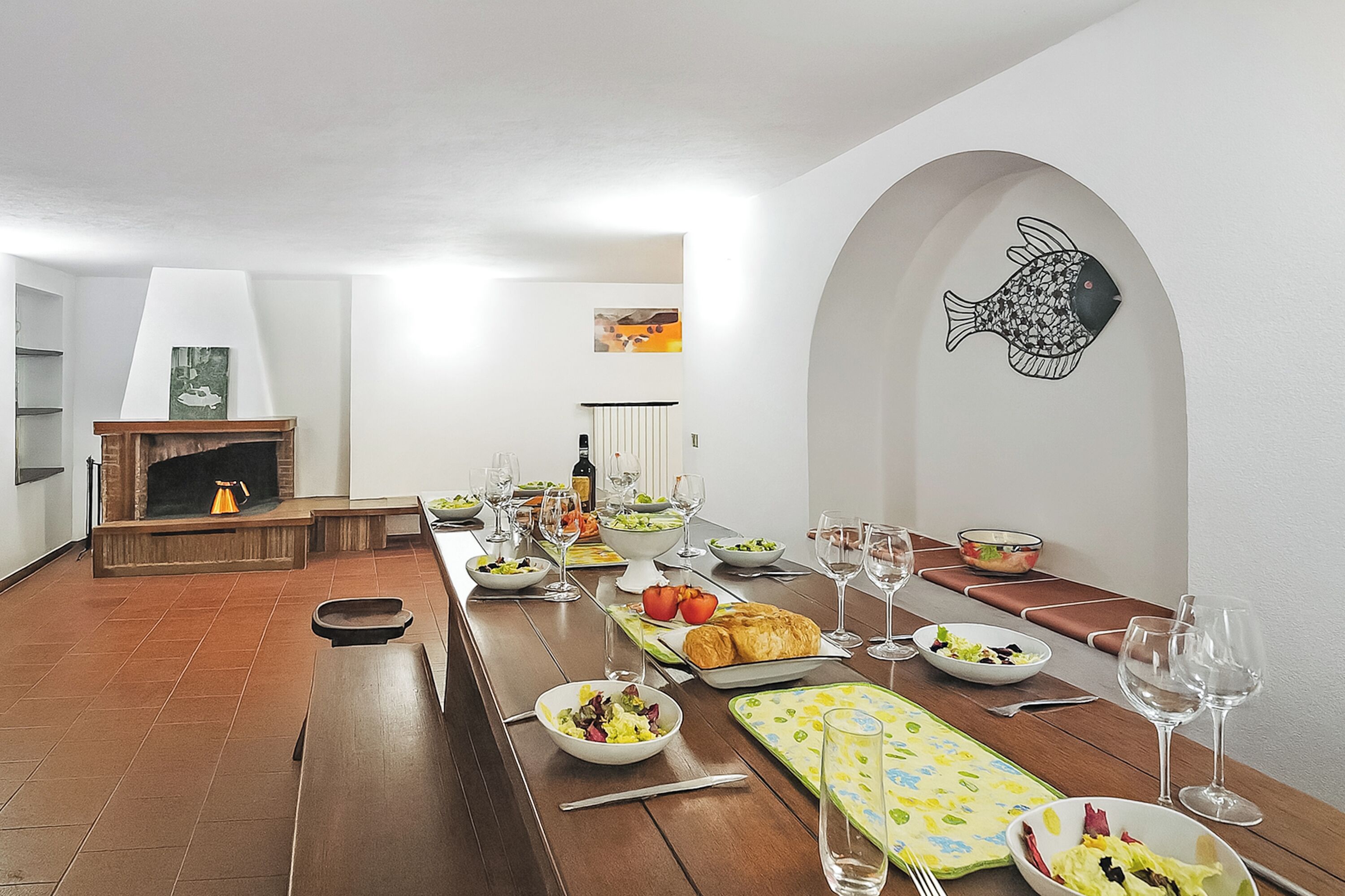 Un comedor con una larga mesa de madera dispuesta para una comida, una chimenea y una obra de arte de un pez colgada en la pared.