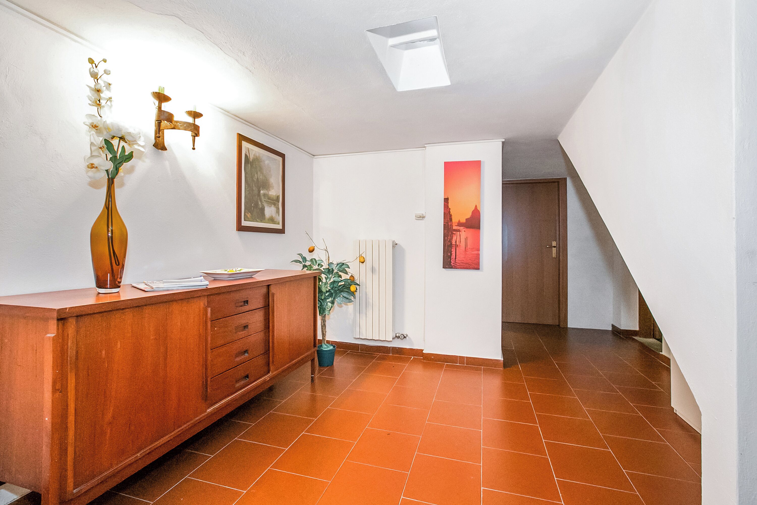 Ein Flur mit einem Holzschrank, einer Blumenvase, einem gerahmten Bild, einer Zimmerpflanze und einer Tür zu einem anderen Raum.