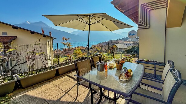 A patio with a table set for two, a large umbrella, and a view of a mountain and sea.