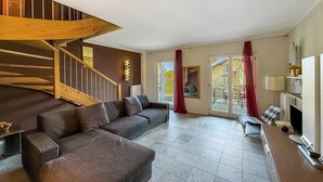 A modern living room with a wooden staircase, a grey sofa, a dining area with a table and chairs, and a view through glass doors to an outdoor patio.