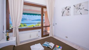 Ein Schlafzimmer mit großem Fenster, einem Bett, einem Nachttisch mit Flasche und Glas sowie zwei gerahmten Bildern an der Wand.