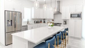 Cozinha moderna com armários brancos, eletrodomésticos de aço inoxidável, uma ilha central com banquetas, iluminação pendente e micro-ondas embutido.