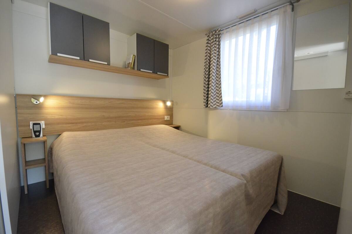 A neatly arranged hotel room with a large bed, a bedside table with a phone, a mirror above the bed, and a window with curtains.