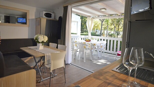 A dining area with a table and chairs, a sofa, a TV, and a balcony with a view of trees. - TopCamp in Union Lido (Cavallino-Treporti)