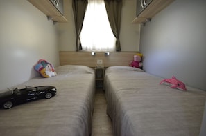 Quarto de hotel pequeno e individual com duas camas, cabeceira de madeira, janela com cortinas, mesa de cabeceira e um carrinho de brinquedo sobre uma das camas.