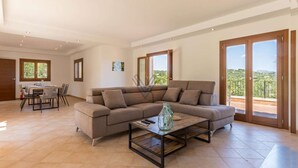 A spacious living area with a large sofa, a coffee table, and a dining table with chairs. There are large windows and a balcony with a view.