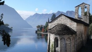 A stone building with a bell tower by a calm lake, surrounded by mountains. - Antico Ulivo (Tremezzina)