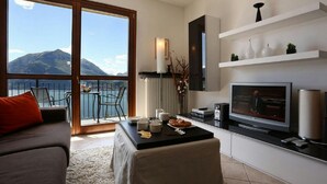 A modern living room with a large TV, a sofa, a coffee table, and a balcony with a view of mountains. - Vista d'Oro Ulivo Apt. 8 (Perledo)