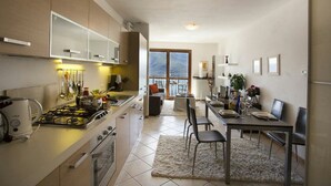 A modern kitchen with a gas stove, oven, and white cabinets. There is a dining area with a table and chairs, and a view of the outdoors through a large window. - Vista d'Oro Ulivo Apt. 8 (Perledo)
