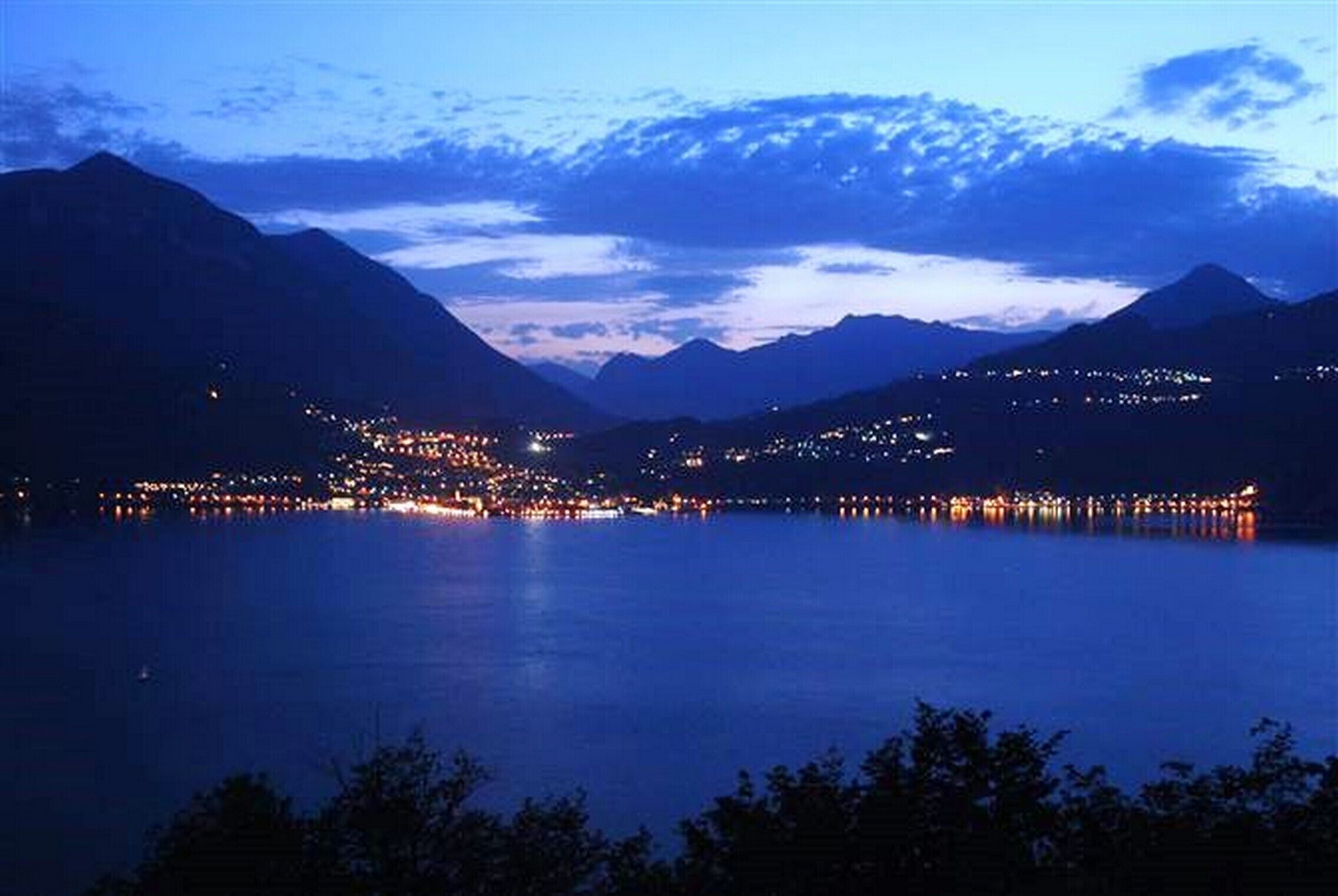 Vista nocturna de un lago rodeado de montañas y un pueblo con edificios iluminados.
