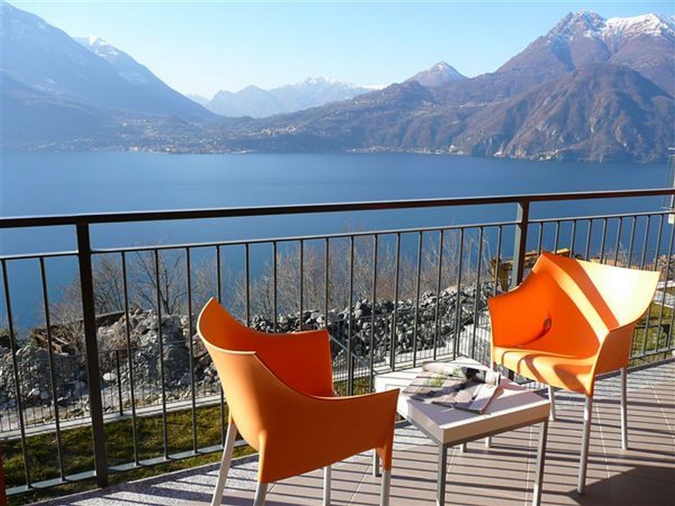 A balcony with two orange chairs and a table, overlooking a lake with mountains in the background.