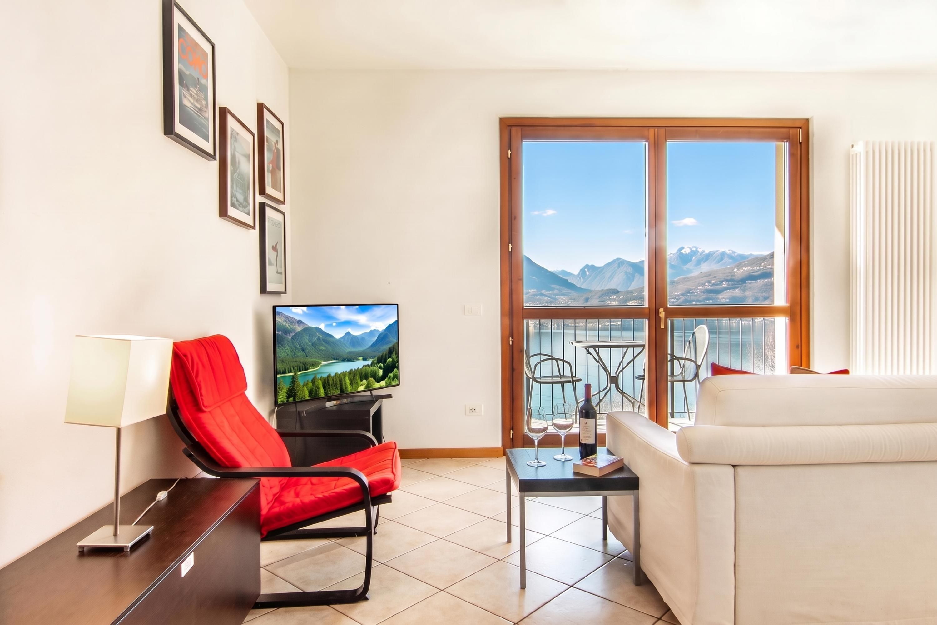 A modern living room with a TV, red chair, sofa, and a balcony with a mountain view.