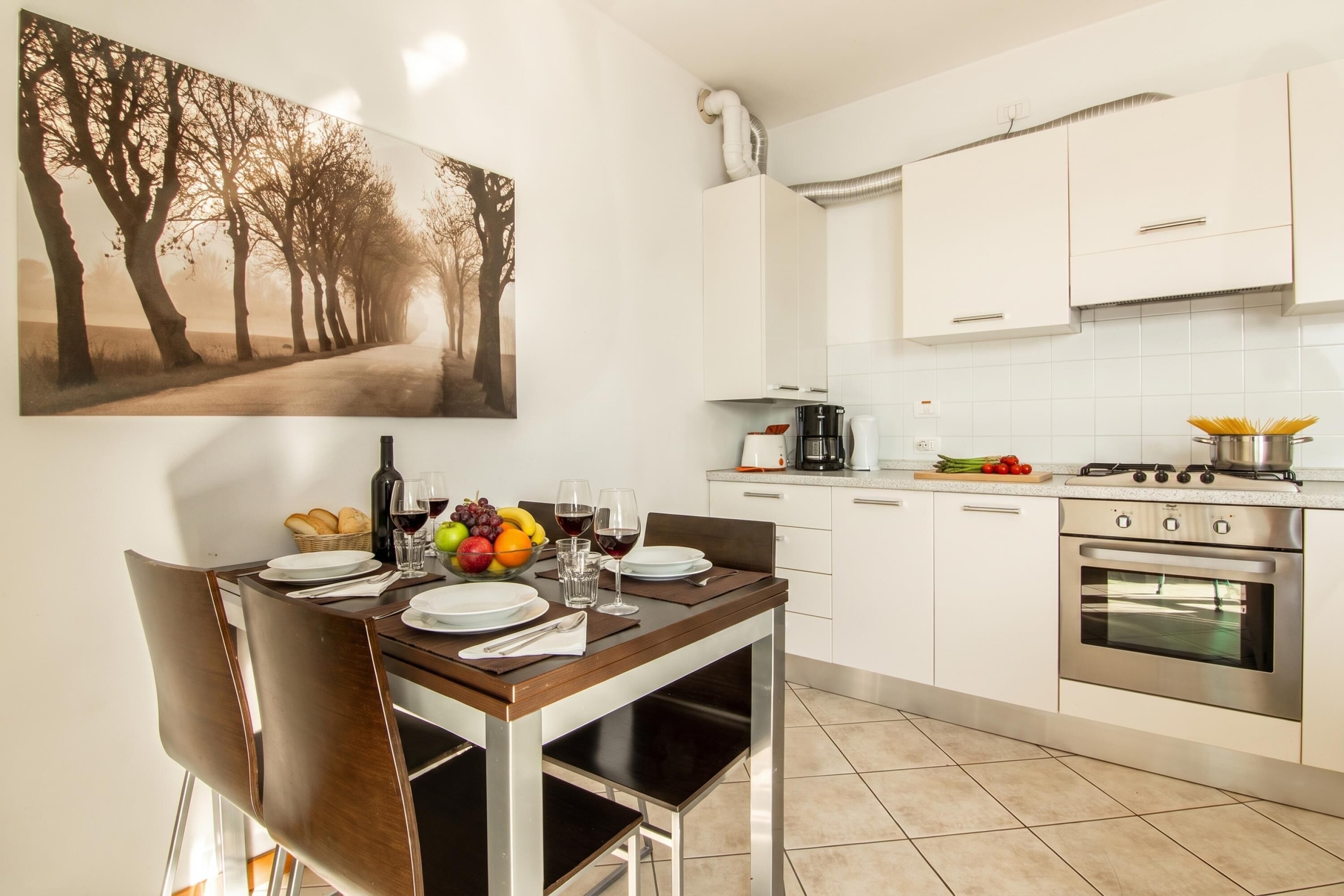 Un comedor con una mesa preparada para dos, una cocina con armarios blancos, una estufa y una cafetera.