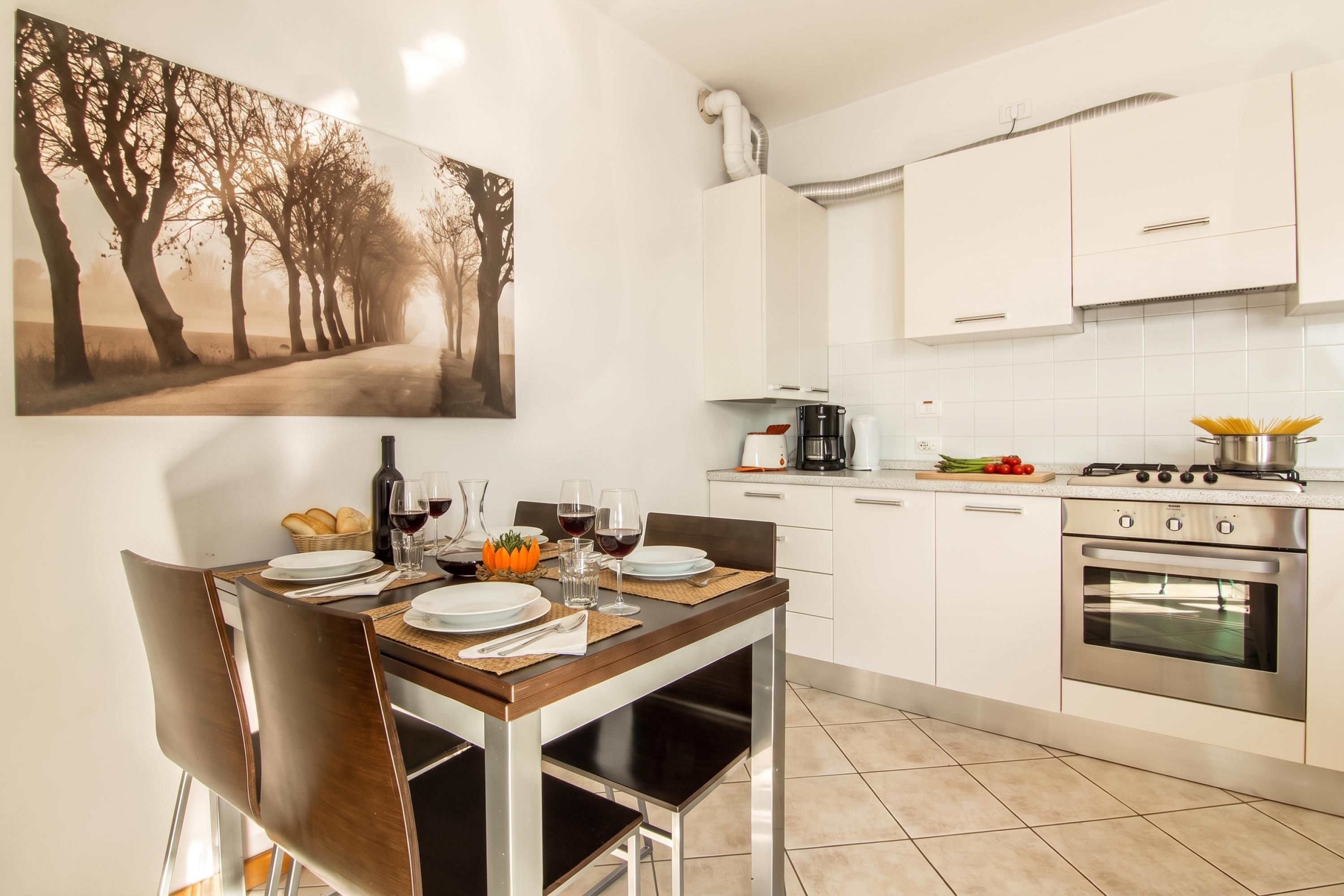 Una cocina moderna con armarios blancos, una mesa de comedor para dos personas y un cuadro de árboles en la pared.