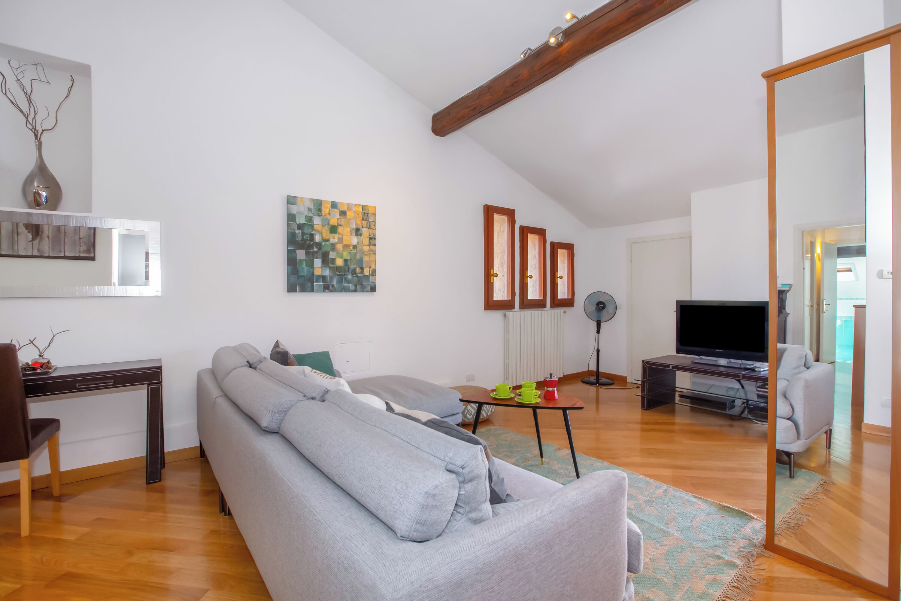 A modern living room with a grey sofa, a wooden coffee table, a TV, and a large mirror.