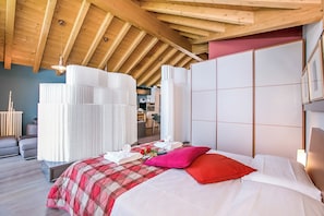 A hotel room with a wooden ceiling, a bed with a plaid bedspread, white linens, a red pillow, and a large window.