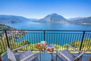 A balcony with two wooden chairs, a small round table, and a tray with a teapot, mug, and flowers. The balcony overlooks a lake with mountains in the background.