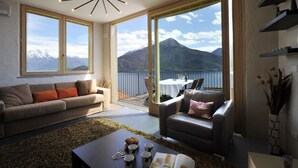 A modern living room with a sofa, armchair, and coffee table. Large windows offer a view of mountains and a lake. - Vista Prima (Pianello del Lario)