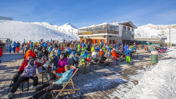 A crowded outdoor seating area with tables and chairs, a building with a balcony, and a snowy mountain backdrop. - Stella Alpina (Livigno)