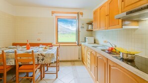 A kitchen with wooden cabinets, a dining table with chairs, a window with a view, and a sink with a pot of spaghetti. - Stella Alpina (Livigno)