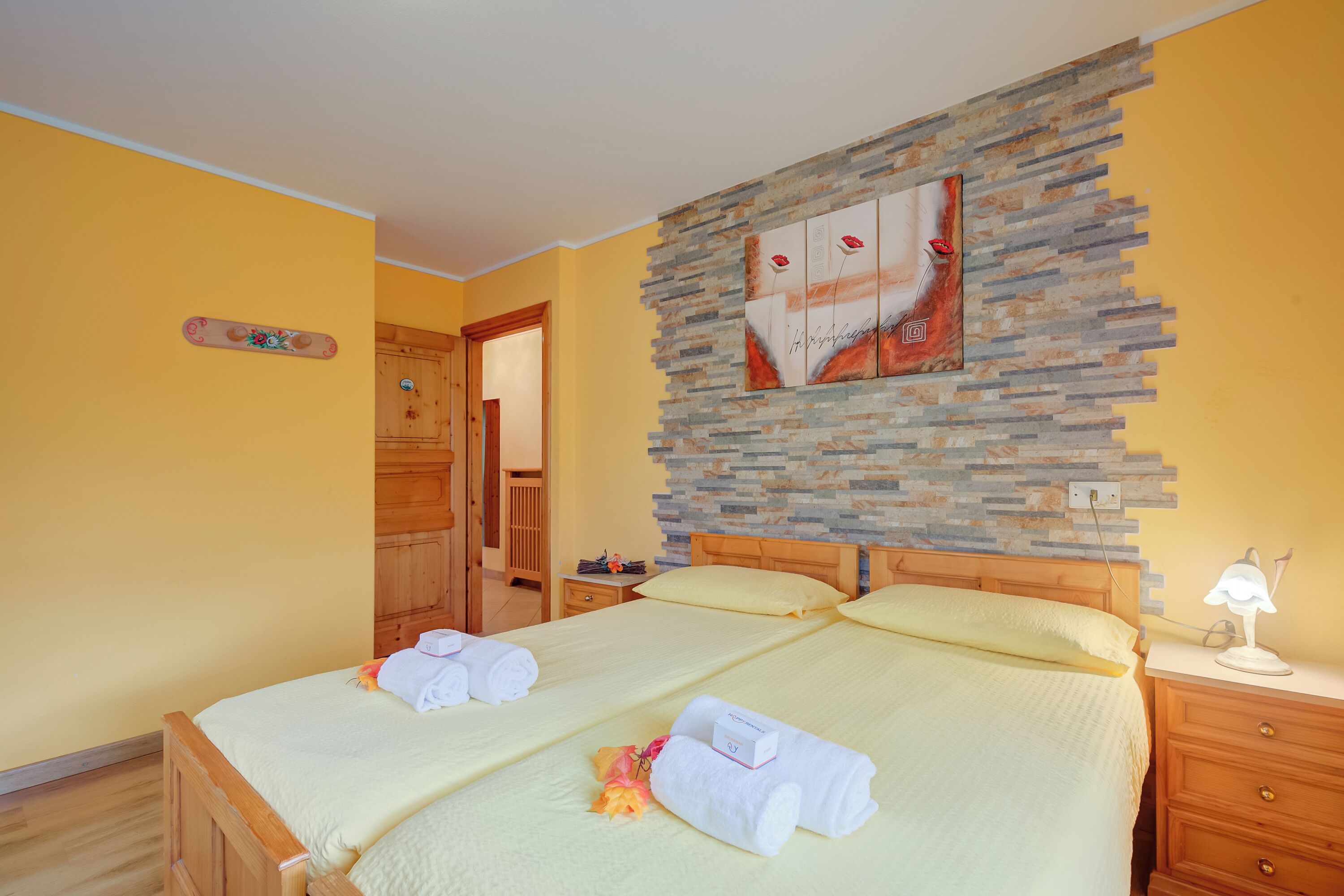 A hotel room with a bed, wooden headboard, a stone accent wall, a painting, a bedside table with a lamp, and a wooden wardrobe.
