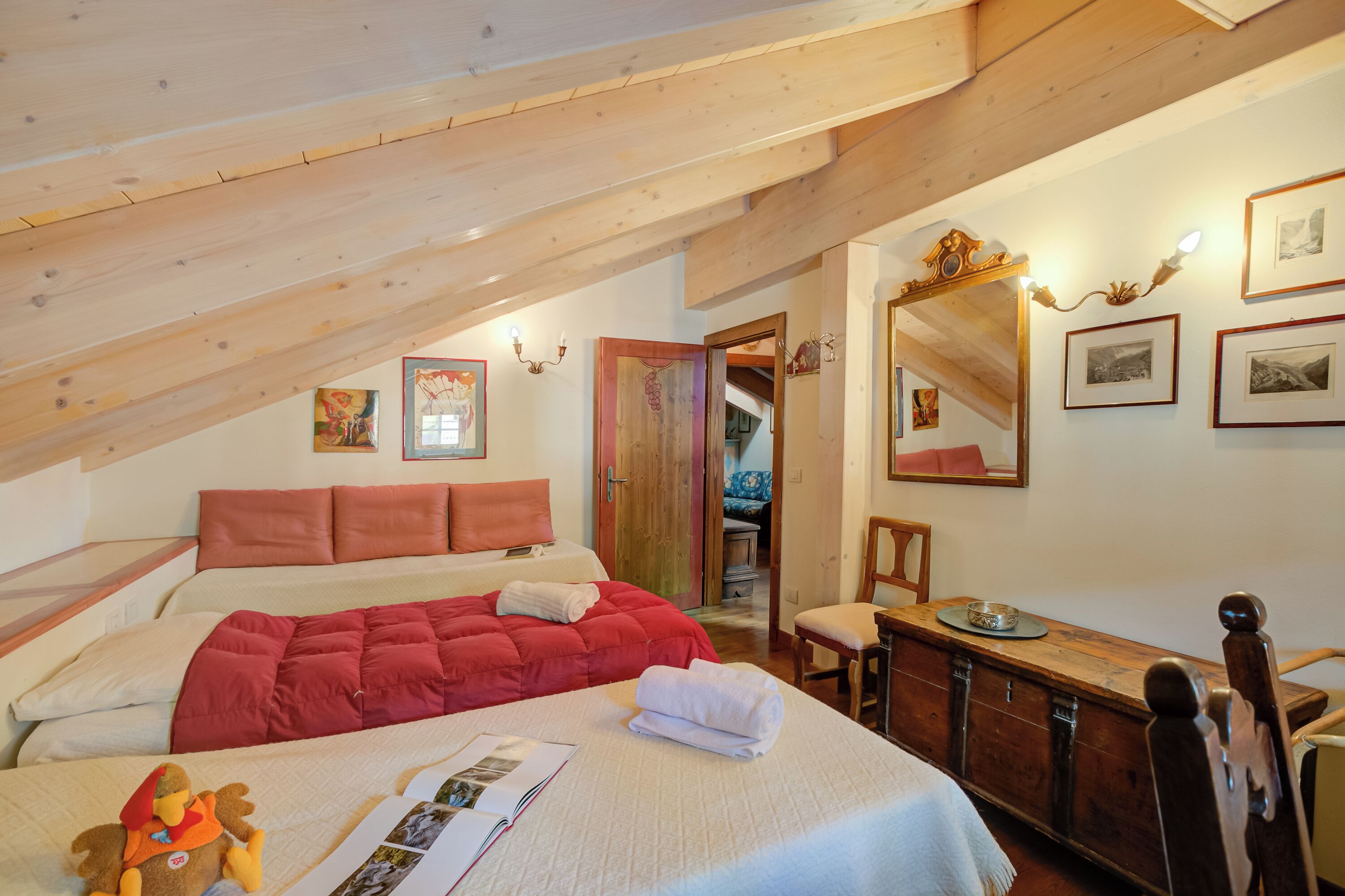 Een knus slaapkamer met schuine wand, een groot bed met rode beddengoed, een houten dressoir en een spiegel aan de muur.