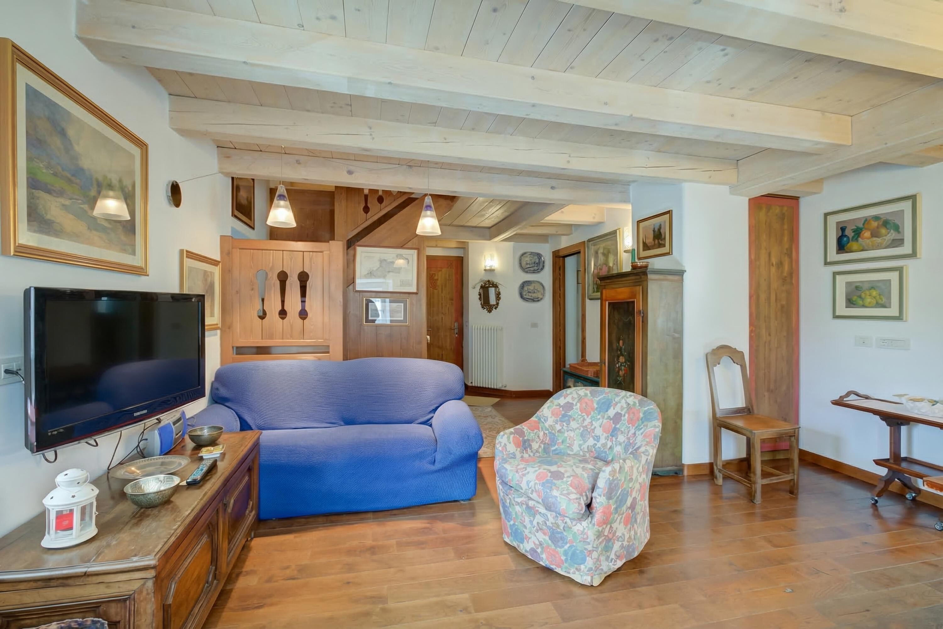 Een woonkamer met een blauwe bank, een fauteuil met bloemenprint, een houten kast, een televisie en kunstwerken aan de muur.
