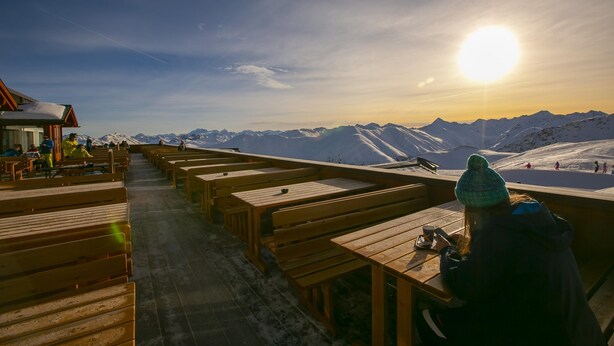 Un pontile in legno con panchine che si affaccia su un paesaggio montuoso innevato al tramonto.