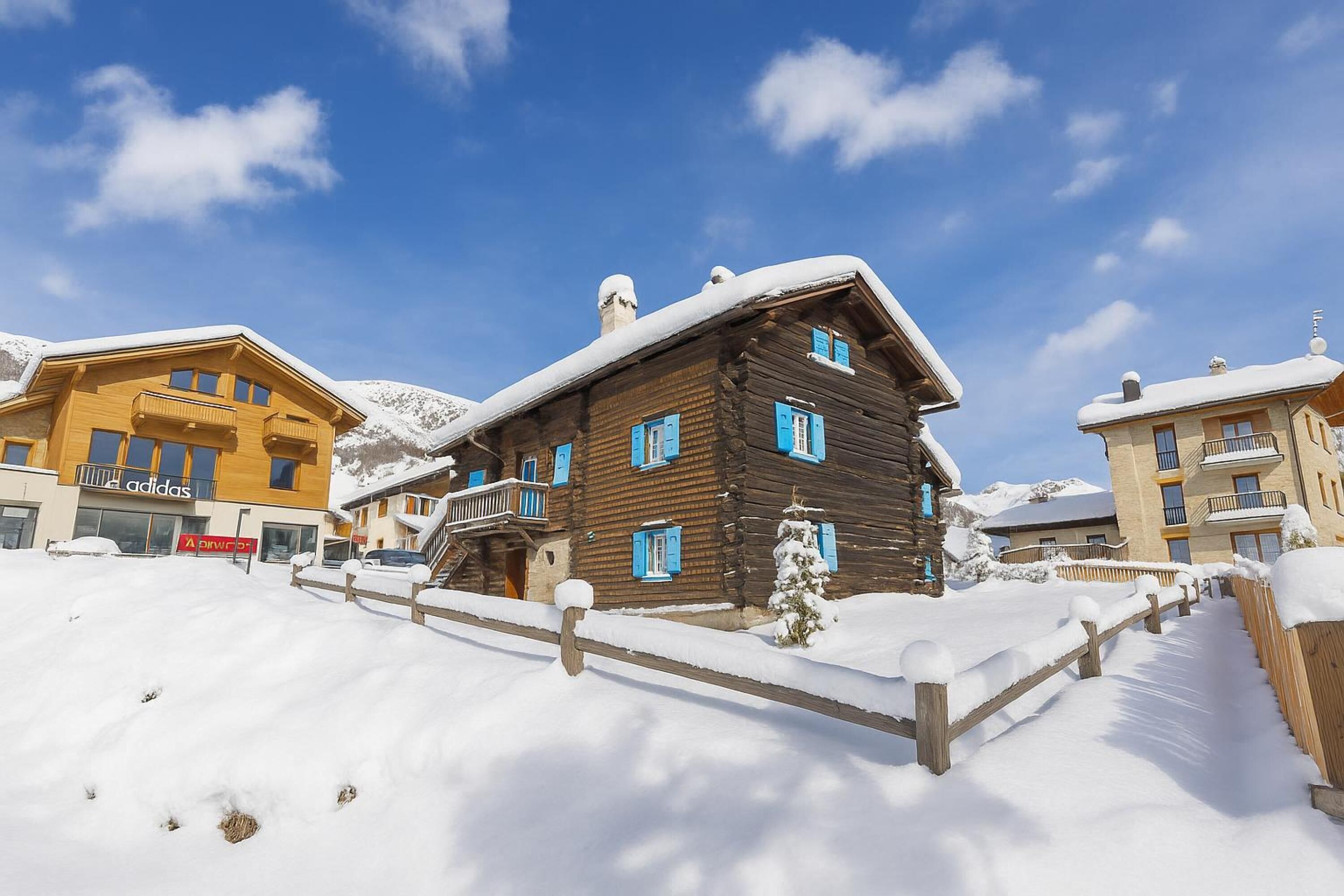 A wooden cabin with a balcony, surrounded by snow-covered ground and buildings.