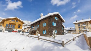 Una baita di legno con un balcone, circondata da terreno innevato e edifici.