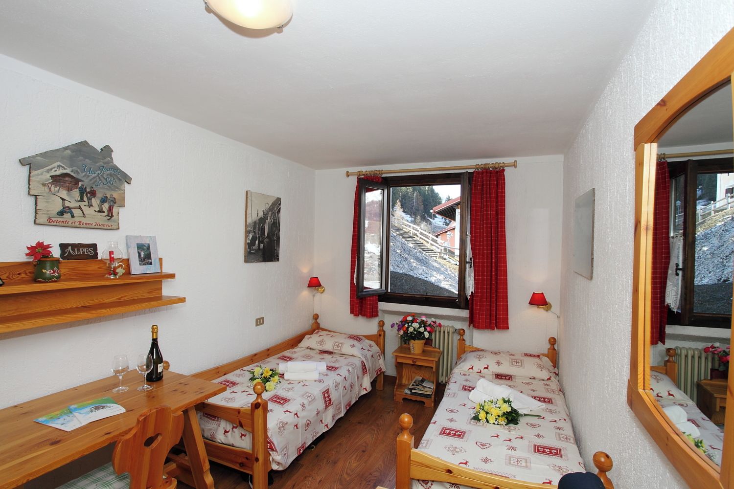 A room with two single beds, a wooden desk, a mirror, and a window with a view of a snowy landscape.