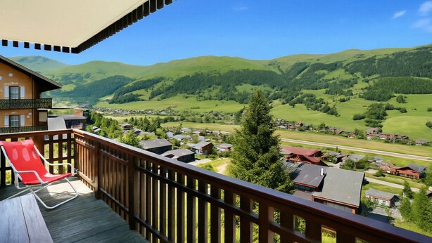 A balcony with a wooden railing, a red chair, and a view of a village with green hills in the background. - Vista Livigno (Livigno)