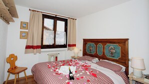 A bedroom with a checkered bedspread, a wooden headboard with floral artwork, a wooden chair, a radiator, and a window with curtains. - Vista Livigno (Livigno)