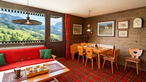 A room with a red plaid floor, a wooden dining table with chairs, a red sofa with green cushions, and a view of green hills through large windows. - Vista Livigno (Livigno)