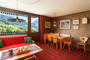 Una stanza con pavimento a quadri rossi, un tavolo da pranzo in legno con sedie, un divano rosso con cuscini verdi e vista su dolci colline attraverso ampie finestre.
