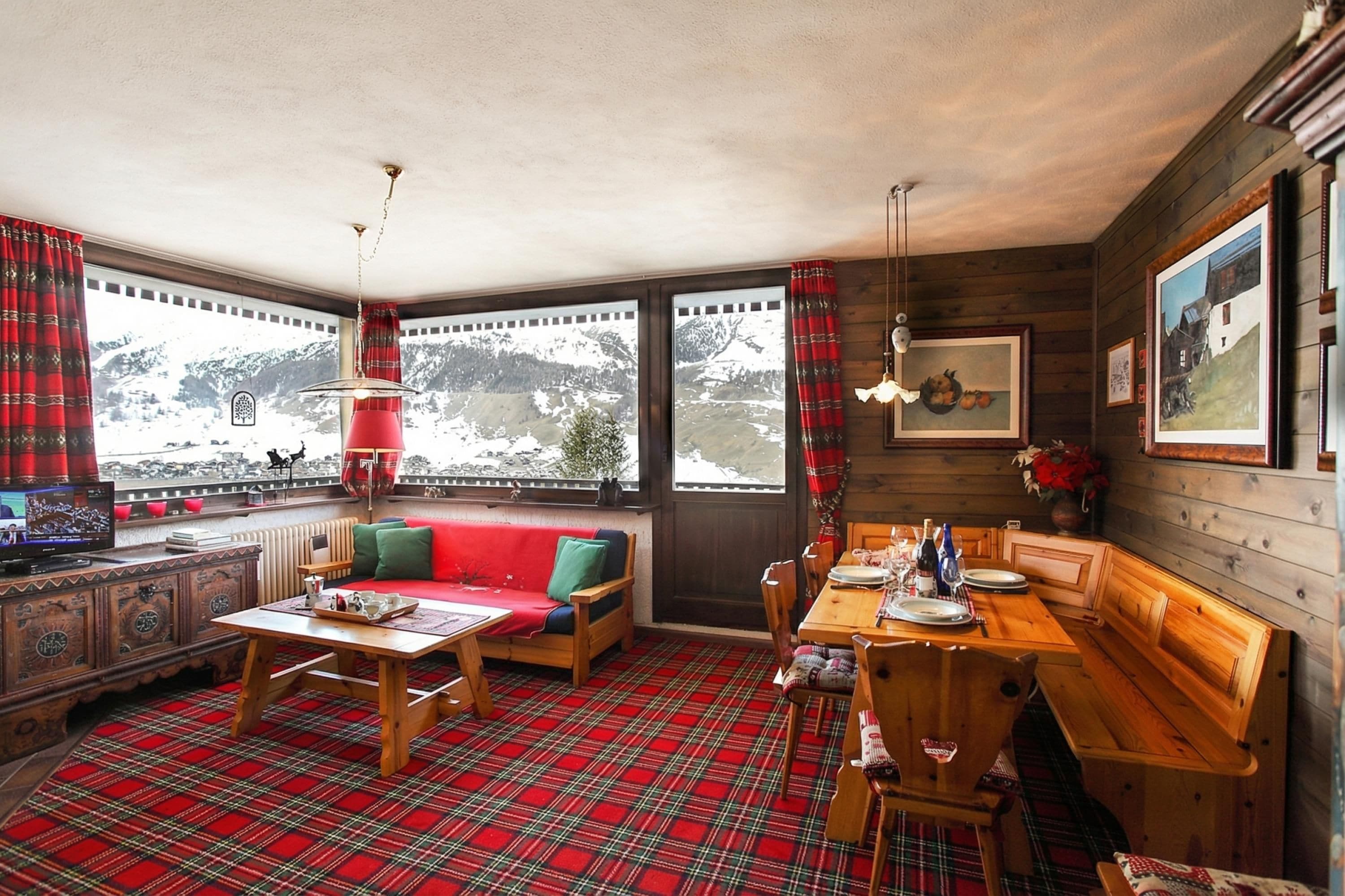 A cozy room with a red plaid carpet, wooden furniture, a dining table set for two, and a large window with curtains.