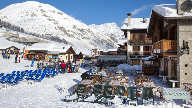 A snow-covered outdoor seating area with wooden tables and chairs, surrounded by buildings with balconies and a mountain backdrop. - Italia 2 Ski in-Ski out 50m (Livigno)