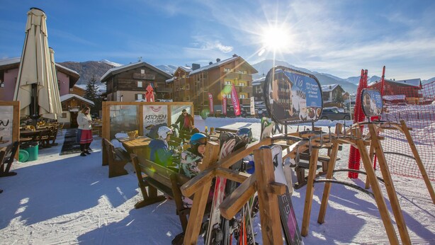 A ski resort with wooden buildings, ski equipment, and a sunlit snowy landscape.