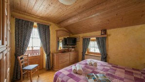 A room with wooden ceiling, a bed with a purple checkered bedspread, a chair, a dresser, a television, and curtains.