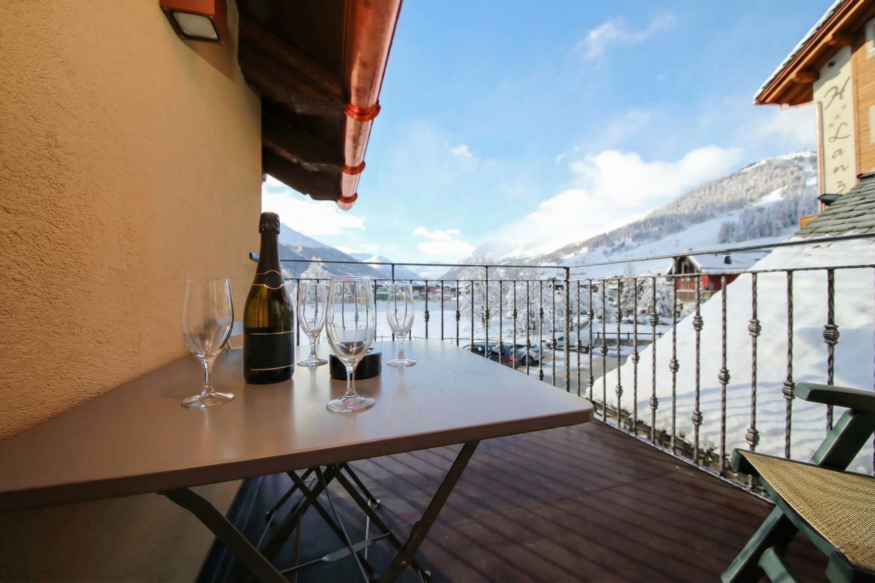 A balcony with a table, wine bottle, and glasses, overlooking a snowy landscape with mountains.