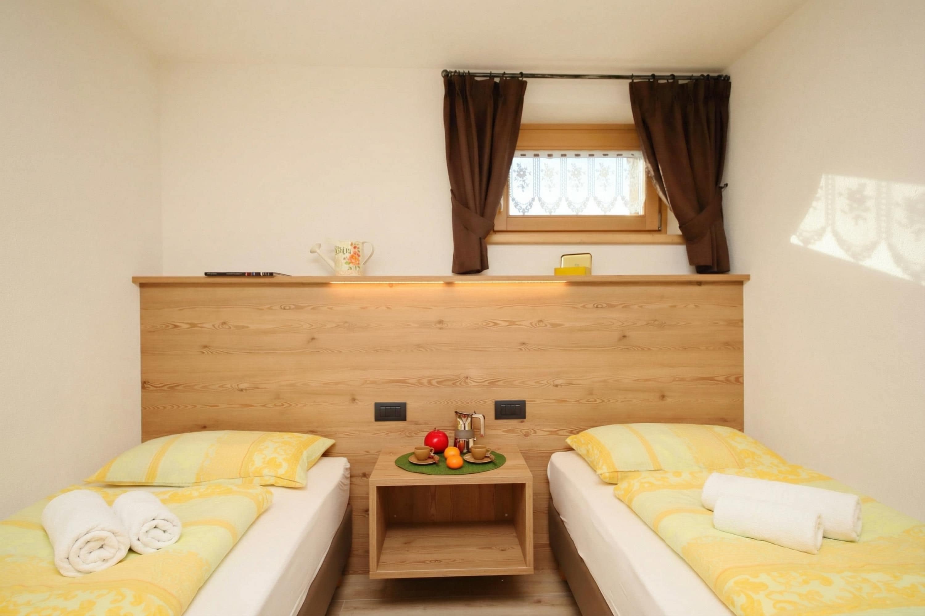 Une chambre avec deux lits, une tête de lit en bois, une petite table sur laquelle se trouve un panier de fruits, et une fenêtre avec des rideaux.