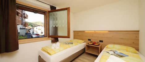 Une chambre d’hôtel avec deux lits, une tête de lit en bois, une fenêtre avec des rideaux et une petite table en bois sur laquelle se trouvent des fruits.