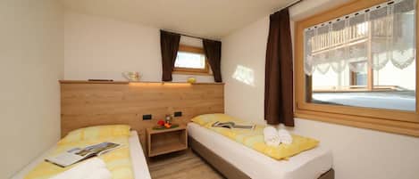 A small, well-lit room with two beds, a wooden headboard, a window with curtains, and a bedside table with a book and a towel.