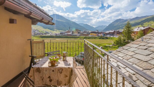 A balcony with a table set for dining, overlooking a mountainous landscape with buildings in the distance.