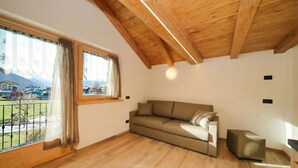 A room with a wooden ceiling, a sofa, a small ottoman, and a large window with curtains.