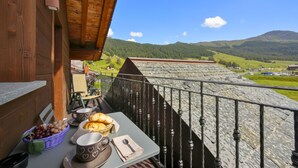 A balcony with a table set for breakfast, a basket of bread, and a scenic view of green hills and a mountain.