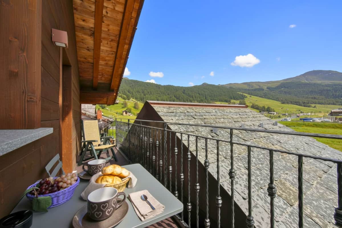A balcony with a table set for breakfast, a basket of bread, and a scenic view of green hills and a mountain.