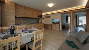 A modern kitchen with wooden cabinets, a dining table set for two, and a living area with a sofa and a television.